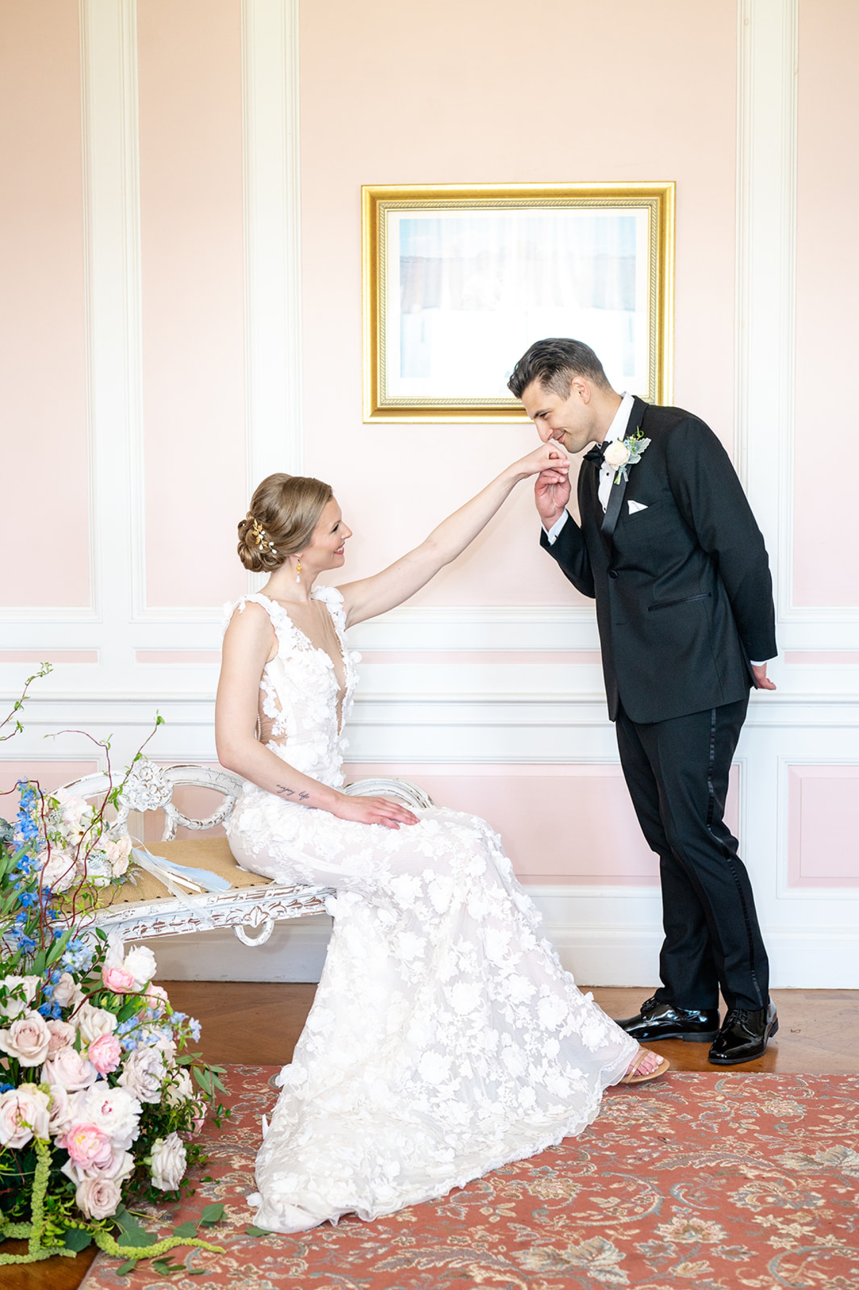 A groom kisses the hand of his bride as she sits on a bench indoors