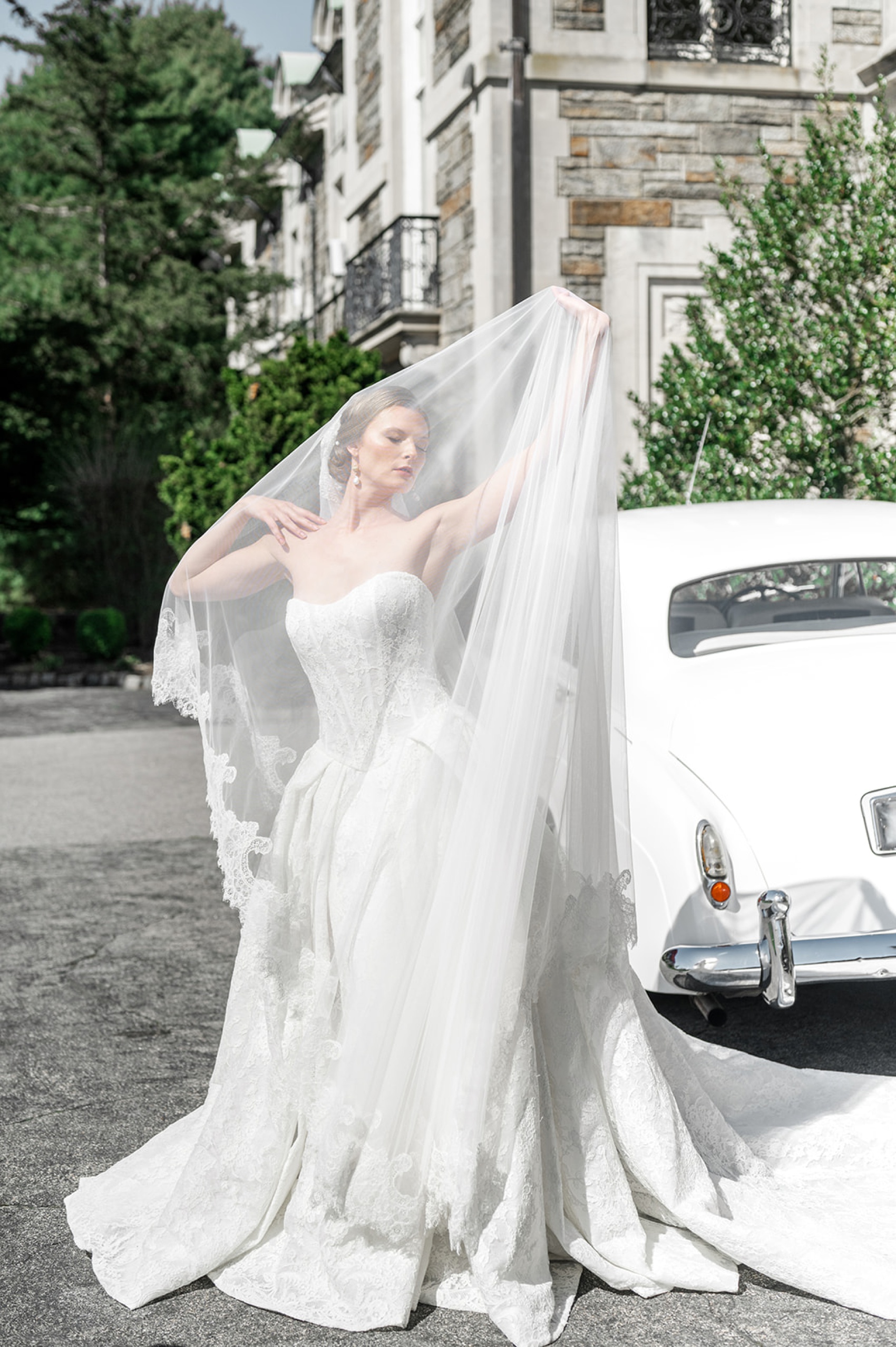 A bride dances under her lace lined veil next to a vintage white car outside the aldrich mansion wedding venue