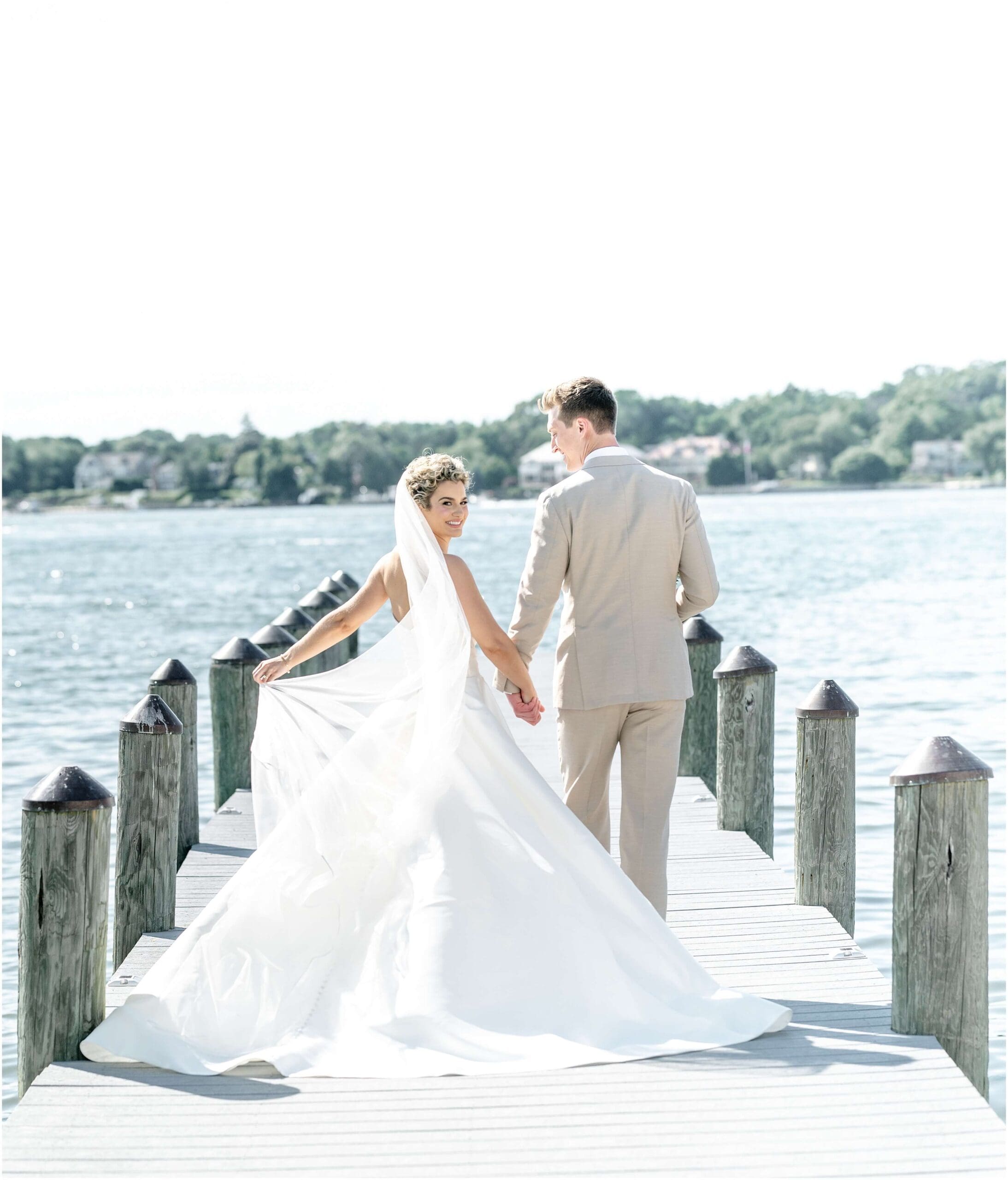 a bride smiles over her shoulder while walking down a dock holding hands with her groom in a tan suit at their Clarks Landing Yacht Club wedding