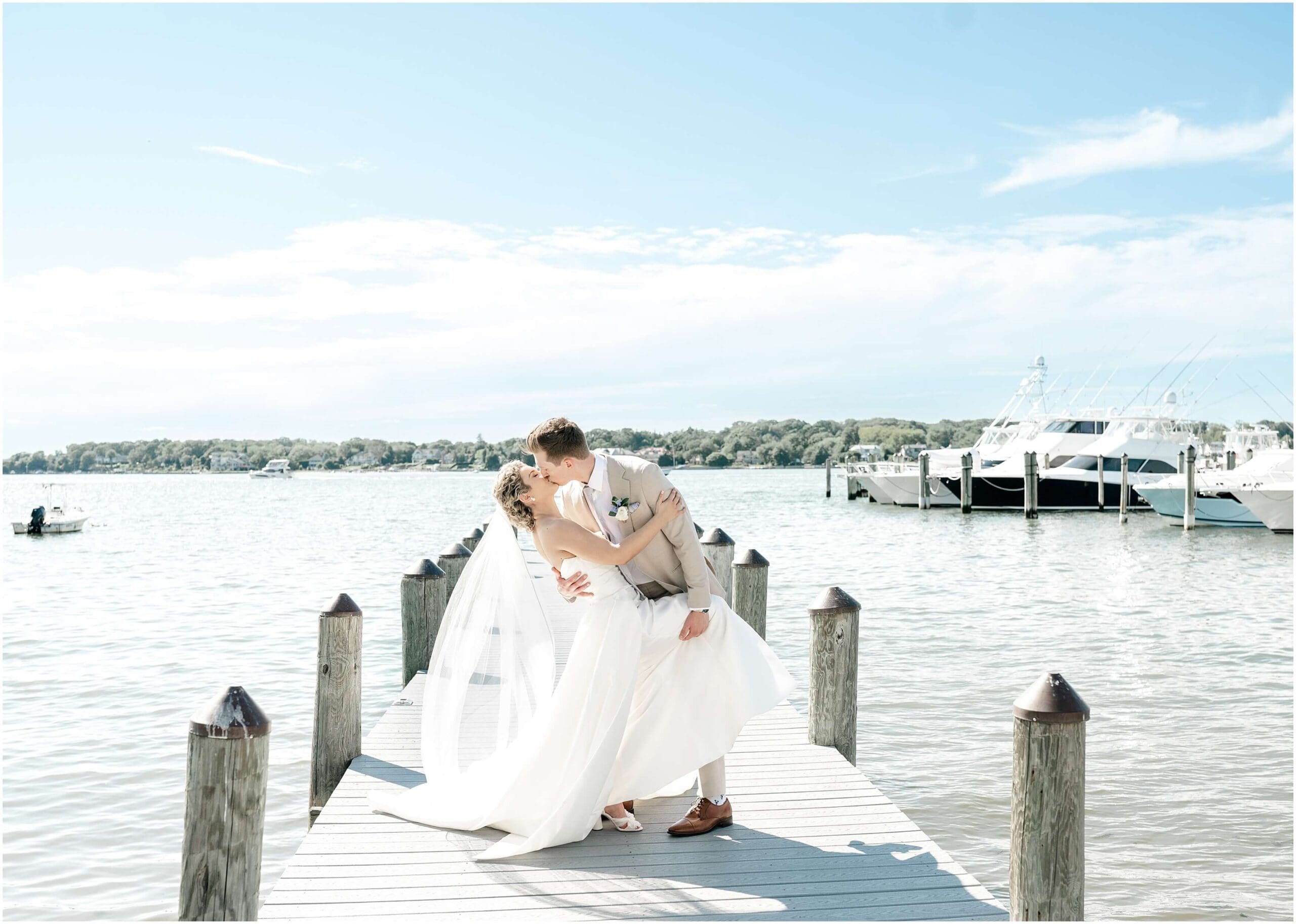 A groom in a tan suit dips his bride while walking on a dock during their Clarks Landing Yacht Club wedding