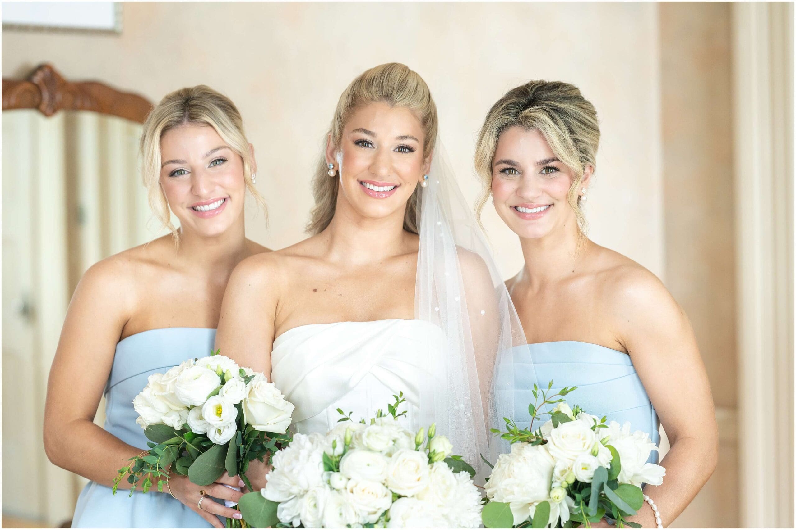 A bride smiles big while standing with her sisters in blue gowns holding white rose bouquets