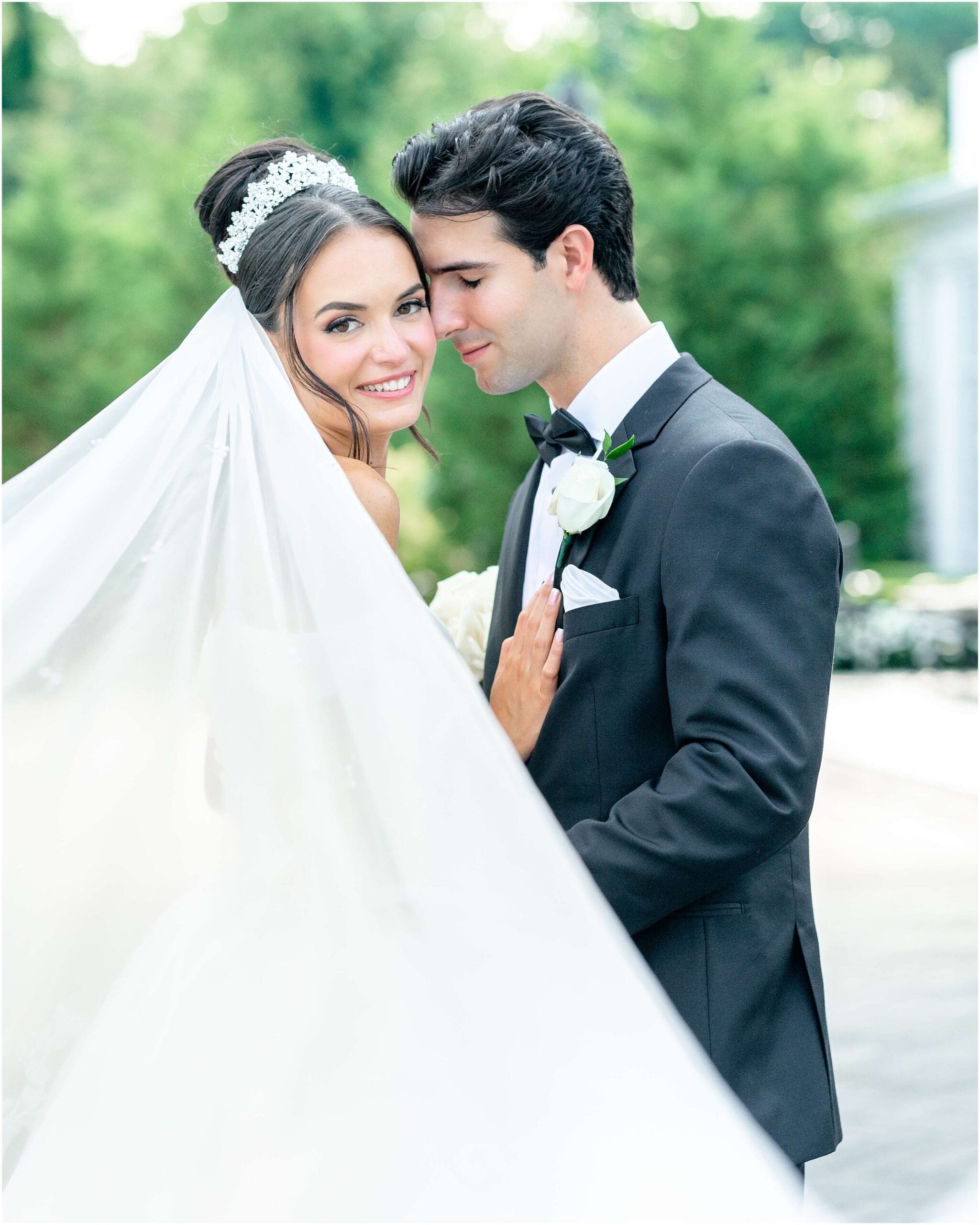 A bride smiles over her shoulder while the groom nuzzles her temple in a black suit while standing in a garden