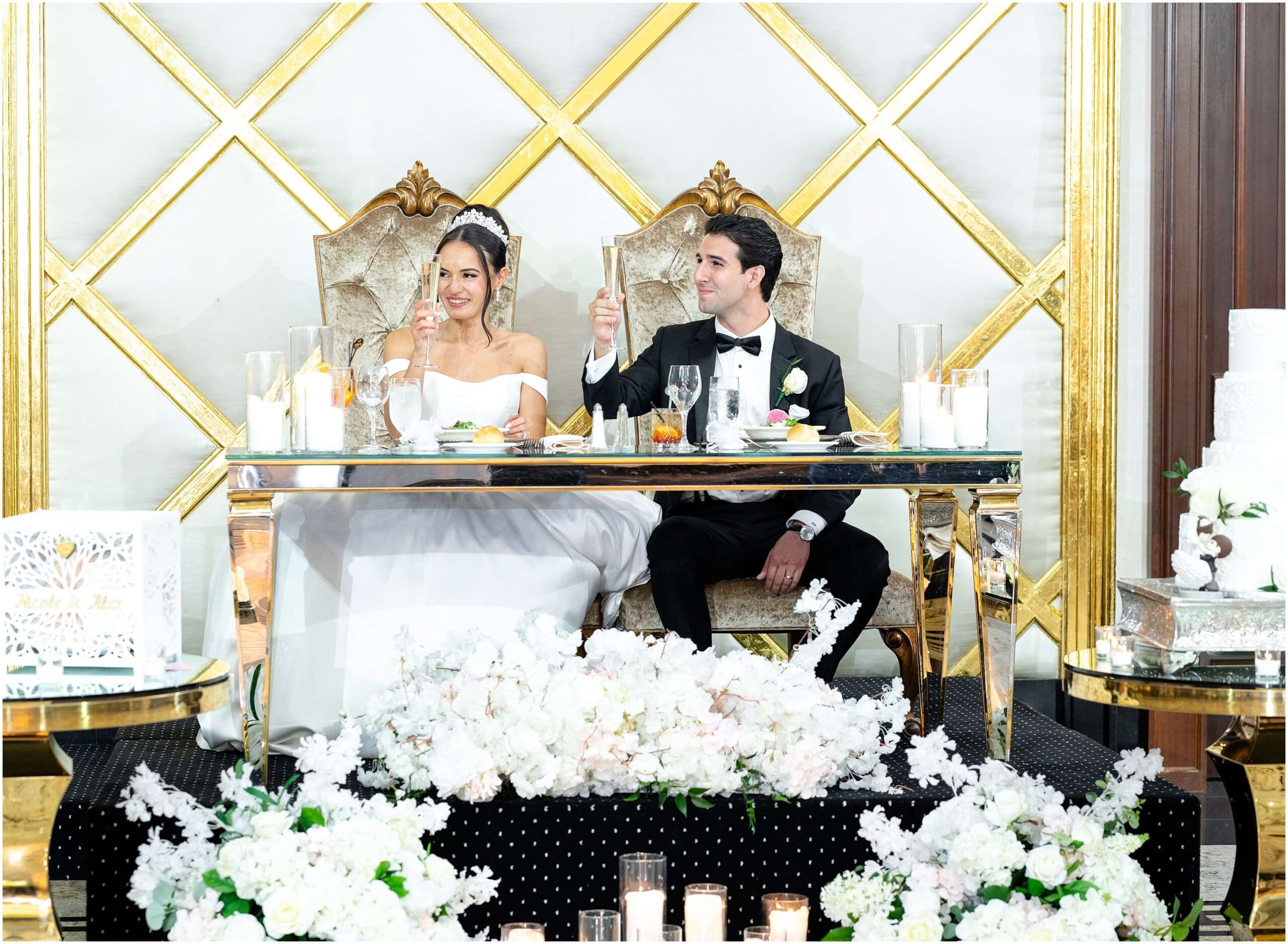 Newlyweds sit at their ornate and flower covered head table on a stage toasting champagne with big smiles during their Shadowbrook at Shrewsbury wedding reception