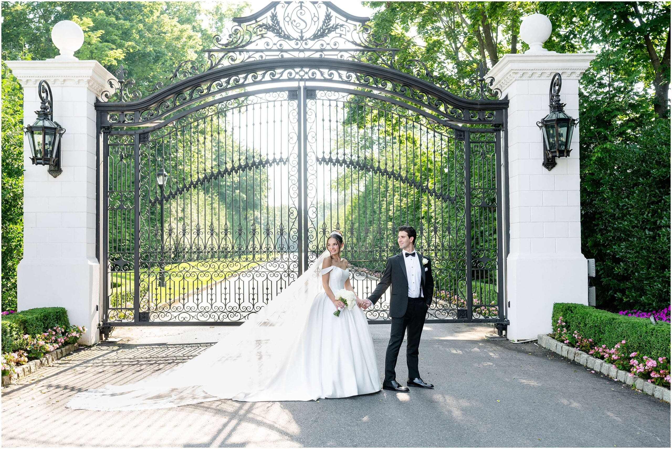 Newlyweds walk holding hands and smiling over their shoulder at the front gates of the Shadowbrook at Shrewsbury wedding venue